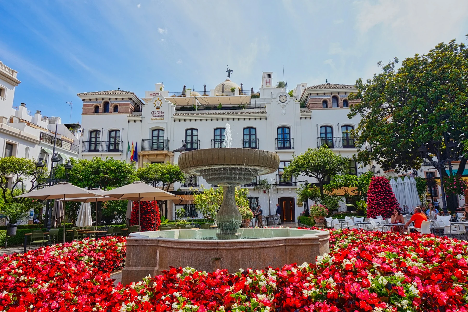 Red flowers, a fountain and café terraces beneath the elegant white façade of El Pilar in Plaza de las Flores, Estepona