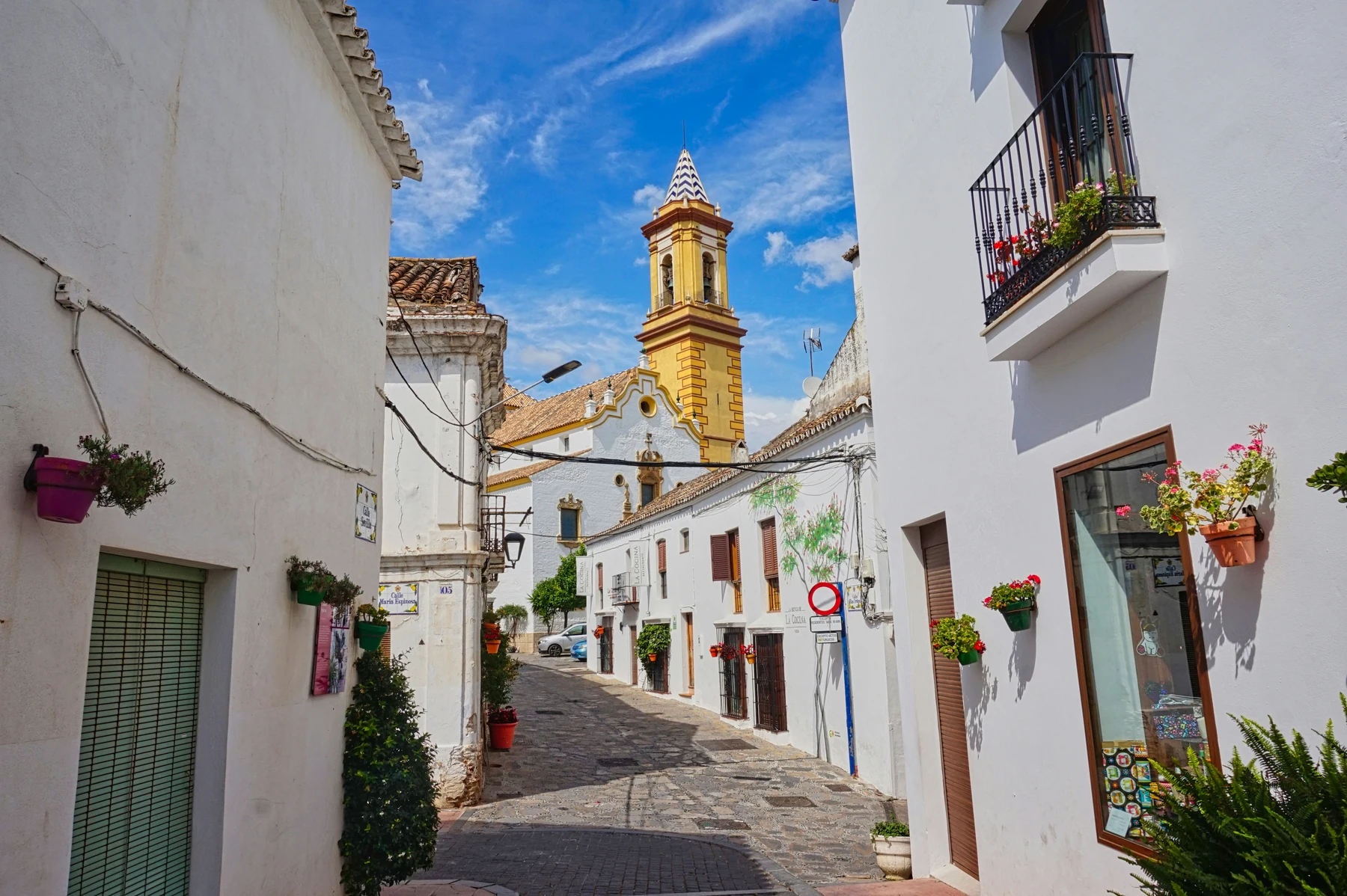 The yellow tower of Nuestra Señora de los Remedios above a flower-lined street in Estepona under a blue sky